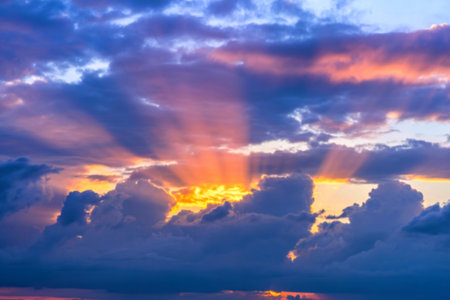 A vibrant sunset scene featuring crepuscular rays piercing through a dramatic, dark cloud formation. The sky showcases a mix of purple, orange, and blue hues.の写真素材