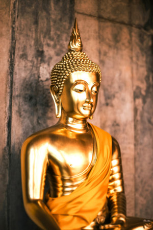 A serene golden Buddha statue sits in meditation, draped in a yellow robe. The statue is beautifully detailed and set against a textured gray background.の素材