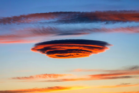 A striking lenticular cloud, illuminated by the setting sun, displays vibrant orange and dark hues against a backdrop of colorful, wispy clouds.の素材