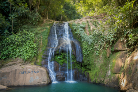 A serene waterfall cascades down moss-covered rocks into a tranquil pool, surrounded by vibrant green tropical foliage. The name Chet Sao Noi is inscribed on a rock.の写真素材