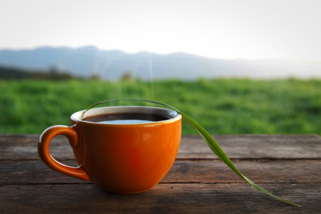 An orange mug filled with steaming coffee sits on a rustic wooden surface. A single green blade of grass rests across the rim, with a blurred green landscape and sky in the background.の写真素材