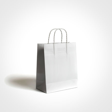 A simple, unbranded white paper shopping bag stands upright on a clean white surface. The bag features a classic design with sturdy, twisted paper handles and a subtle shadow.の写真素材