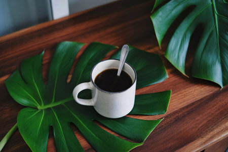 A cup of black coffee sits on a large, vibrant green monstera leaf, resting on a rich brown wooden surface. A spoon is nestled in the cup.の写真素材