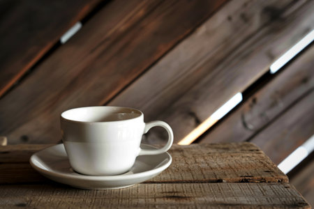 A clean white coffee cup and saucer rests on a weathered wooden table. The background features diagonally arranged wooden planks, creating a rustic and cozy atmosphere.の写真素材