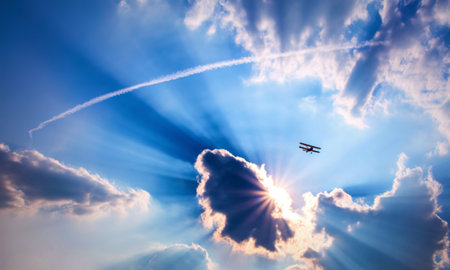 A red biplane is silhouetted against a vibrant blue sky, with sun rays bursting through clouds. A contrail streaks across the upper portion of the image.の写真素材
