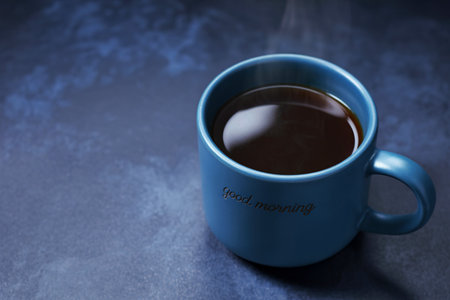 A close-up shot of a blue ceramic mug filled with dark coffee, emitting gentle steam. The mug has 'good morning' inscribed on its side, set against a textured dark background.の写真素材