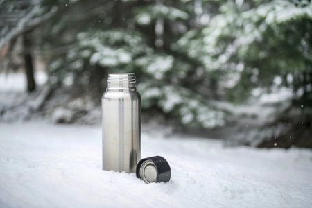 A silver stainless steel thermos bottle and its cap rest on a snowy surface, with a blurred...