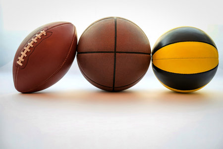 An American football, a rugby ball, and a basketball are displayed side by side on a white surface with a bright background.の写真素材