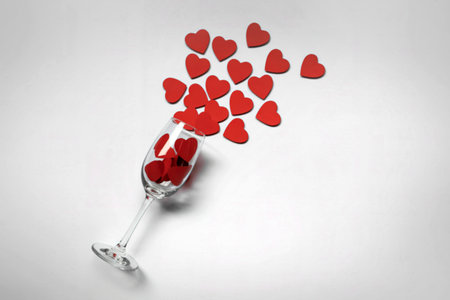 A champagne flute tipped over, spilling a cascade of small red heart shapes onto a white surface.