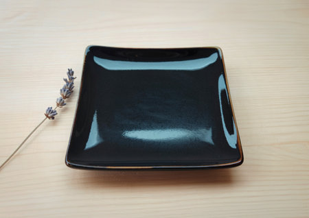 A dark, glossy black square plate sits on a light-colored wooden surface. A single dried lavender...