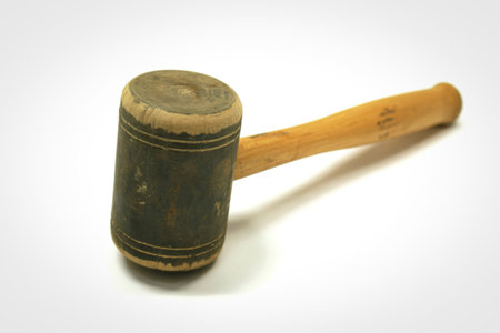 A close-up, angled view of a weathered wooden mallet with a dark, cylindrical head and a smooth,...