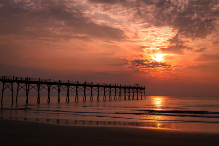 A long pier stretches into the ocean at sunrise, silhouetted against a vibrant orange and pink sky....