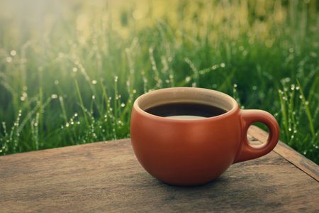 A close-up shot of a warm, brown coffee cup filled with dark coffee sits on a weathered wooden surface. The background features lush green grass sparkling with morning dew.の写真素材