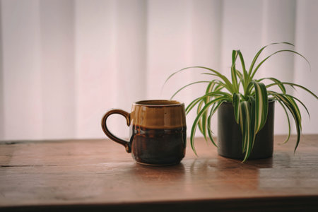 A rustic brown ceramic mug sits beside a spider plant in a dark gray pot on a wooden surface, near a bright window. The scene is simple and calming.の写真素材