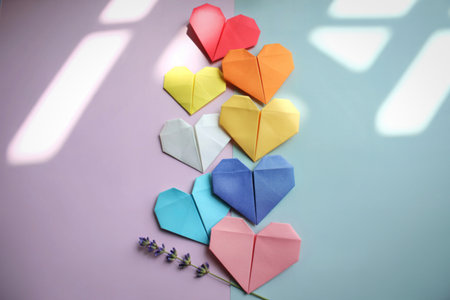 A vertical arrangement of seven colorful origami hearts, casting soft shadows from sunlight filtering through blinds, against a light blue background.の写真素材