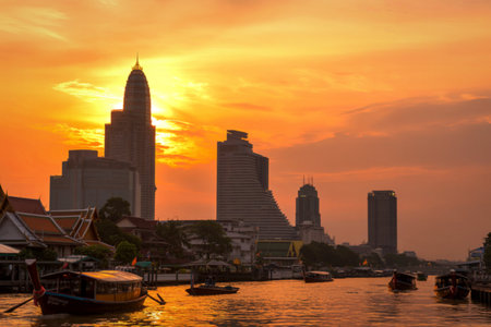 Silhouetted skyscrapers rise against a vibrant orange sunset over the Chao Phraya River, with boats navigating the water below.の写真素材