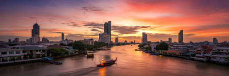 A traditional boat sails down a river towards a modern city skyline at sunset with vibrant orange and pink clouds.の写真素材