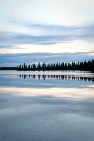 A calm lake mirrors a distant line of dark trees against a soft, overcast sky, creating a peaceful, symmetrical landscape.の写真素材