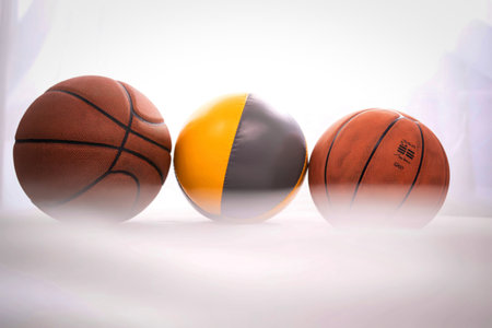 Three spherical objects are lined up on a white surface. Two are traditional basketballs, and one is a unique yellow and gray ball.の写真素材