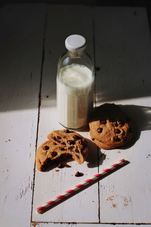 A glass bottle filled with milk sits next to two cookies and a striped straw on a weathered wooden table, bathed in sunlight.の写真素材