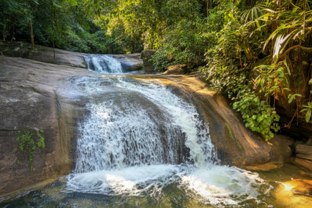 A beautiful cascade of water flows over moss-covered rocks, surrounded by vibrant green foliage and dappled sunlight.の写真素材