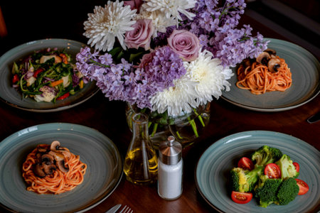 A beautifully arranged table features four individual servings of pasta and salad, complemented by a vibrant bouquet of flowers in a vase.の写真素材
