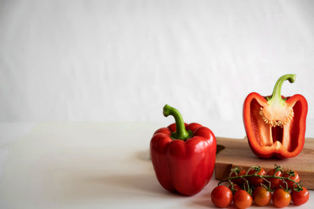 A whole red bell pepper and a halved red bell pepper sit with cherry tomatoes on a wooden cutting board against a white background.の写真素材
