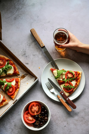 A close-up overhead view of a freshly baked pizza slice on a plate, accompanied by a bowl of tomatoes and olives, and a hand holding a drink.の写真素材