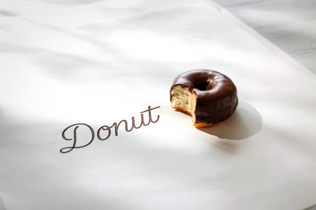 A single, perfectly glazed chocolate donut sits on a white surface, with a bite revealing its soft interior. The word 'Donut' is written in elegant script.の写真素材