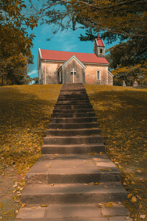A long stone pathway leads up a grassy hill towards a quaint stone church with a red roof, surrounded by colorful autumn foliage.の写真素材