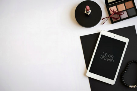 A modern flat lay featuring a smartphone, eyeshadow palette, and jewelry, neatly arranged on a white surface with black paper.の写真素材