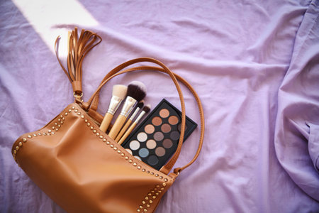A stylish gold handbag spills open revealing makeup brushes and a neutral eyeshadow palette, artfully arranged on a soft, draped fabric.の写真素材