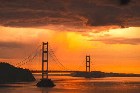 The silhouette of a suspension bridge stands against a fiery orange and yellow sunset sky with dramatic clouds.の写真素材