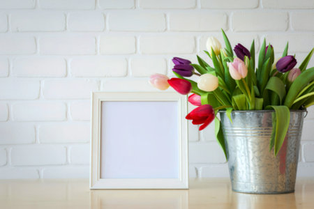 A vibrant bouquet of multi-colored tulips in a metal bucket sits next to a blank white picture frame...