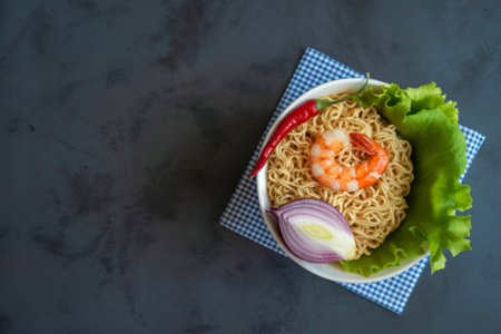 A top-down view of a bowl of instant noodles topped with a shrimp, red chili, red onion, and lettuce, presented on a patterned napkin.の写真素材