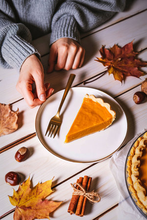 A person in a gray sweater reaches for a slice of pumpkin pie on a white plate, surrounded by fall leaves and cinnamon sticks.の写真素材