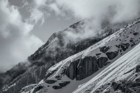 A black and white photograph capturing a rugged mountain landscape covered in snow. The image...