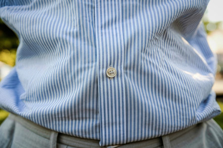 A close-up image of a striped shirt featuring a button on the collar. The shirt has vertical blue...