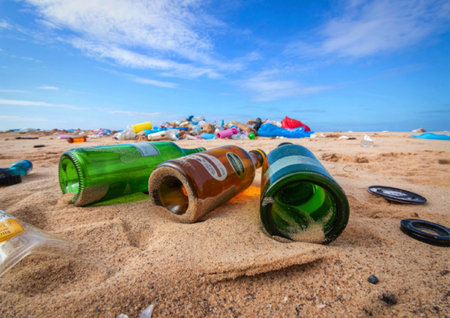 An image of a sandy beach littered with empty bottles and various pieces of trash under a clear blue...