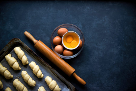 A baking tray with rolled dough, a wooden rolling pin, and a bowl of eggs on a dark surface.