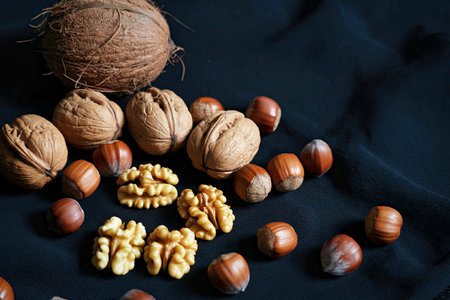 A collection of various nuts, including walnuts, hazelnuts, and a coconut, arranged on a dark...