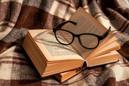 An open book with eyeglasses resting on top, placed on a cozy plaid blanket. The scene evokes a...