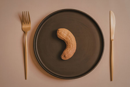 A solitary cashew nut is centered on a dark plate, flanked by a gold fork and knife, set against a beige background.の写真素材