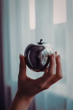 A hand holding a reflective apple, with a blurred background of a window with white curtains.の写真素材
