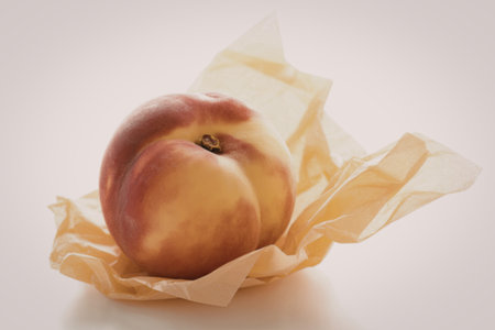 A ripe peach with red and yellow skin is placed on crumpled yellow tissue paper, set against a clean white background.の写真素材