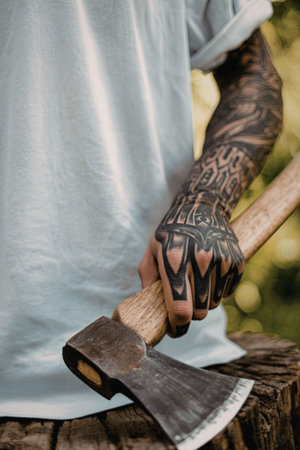 A man with a heavily tattooed arm is holding an axe on a tree stump, suggesting a scene of outdoor work or activity.の写真素材