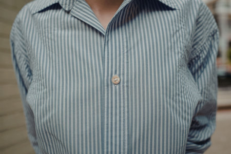 A person is shown wearing a blue and white striped shirt with a buttoned collar, standing outside with a blurred background.の写真素材