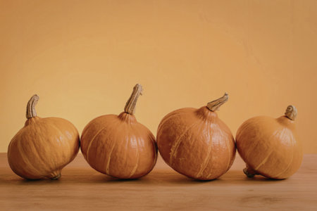 The image features four small, orange pumpkins lined up on a wooden table or surface, set against a bright orange background that gradates to a lighter shade towards the center. The pumpkins are all roughly the same size and have a similar shape, with visible stems and subtle ridging on their surface.の写真素材