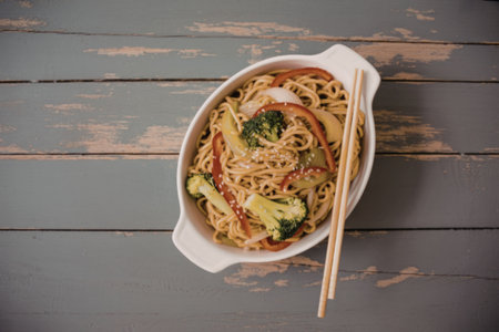 A bowl of noodles mixed with broccoli, red peppers, and onions, served with chopsticks on a distressed gray wooden table.の写真素材