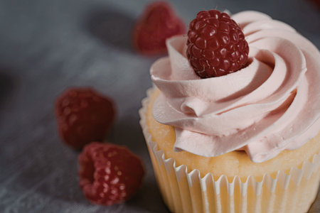 A close-up image of a yellow cupcake with pink raspberry frosting and a fresh raspberry on top, surrounded by scattered raspberries on a gray background.の写真素材
